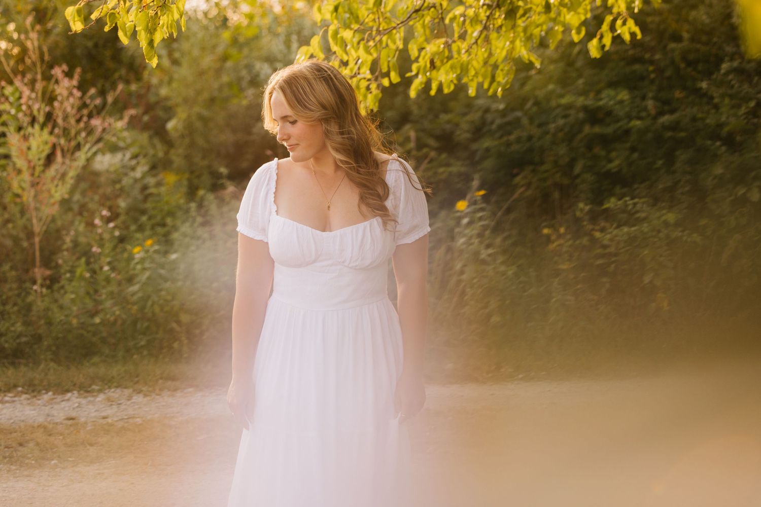 Someone in a flowing white wedding dress poses outdoors during golden hour with yellow foliage in the background.