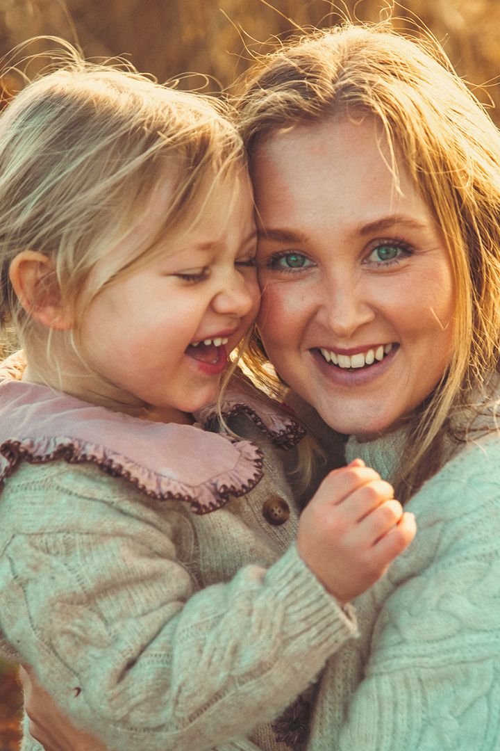 Mor och dotter i varm kvällssol som kramas, jordnära färger, varmt ljus. Golden hour. Flickan skrattar.