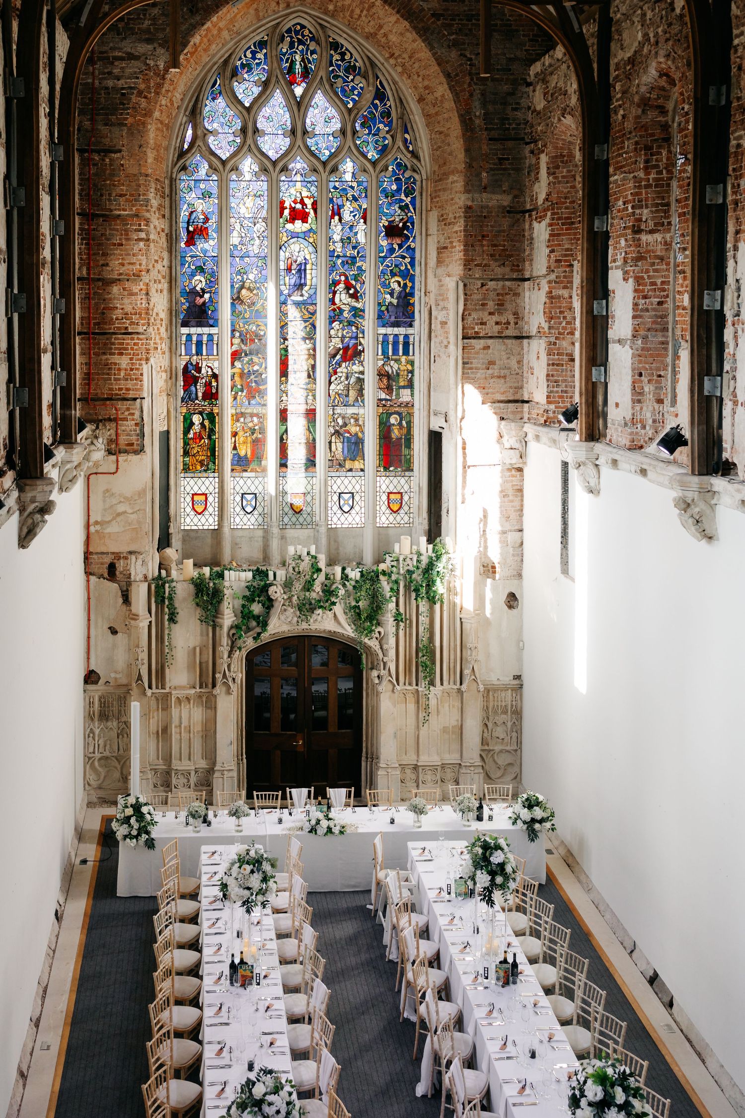 Wedding reception tables arranged in rows beneath a grand stained glass window in a historic church interior.