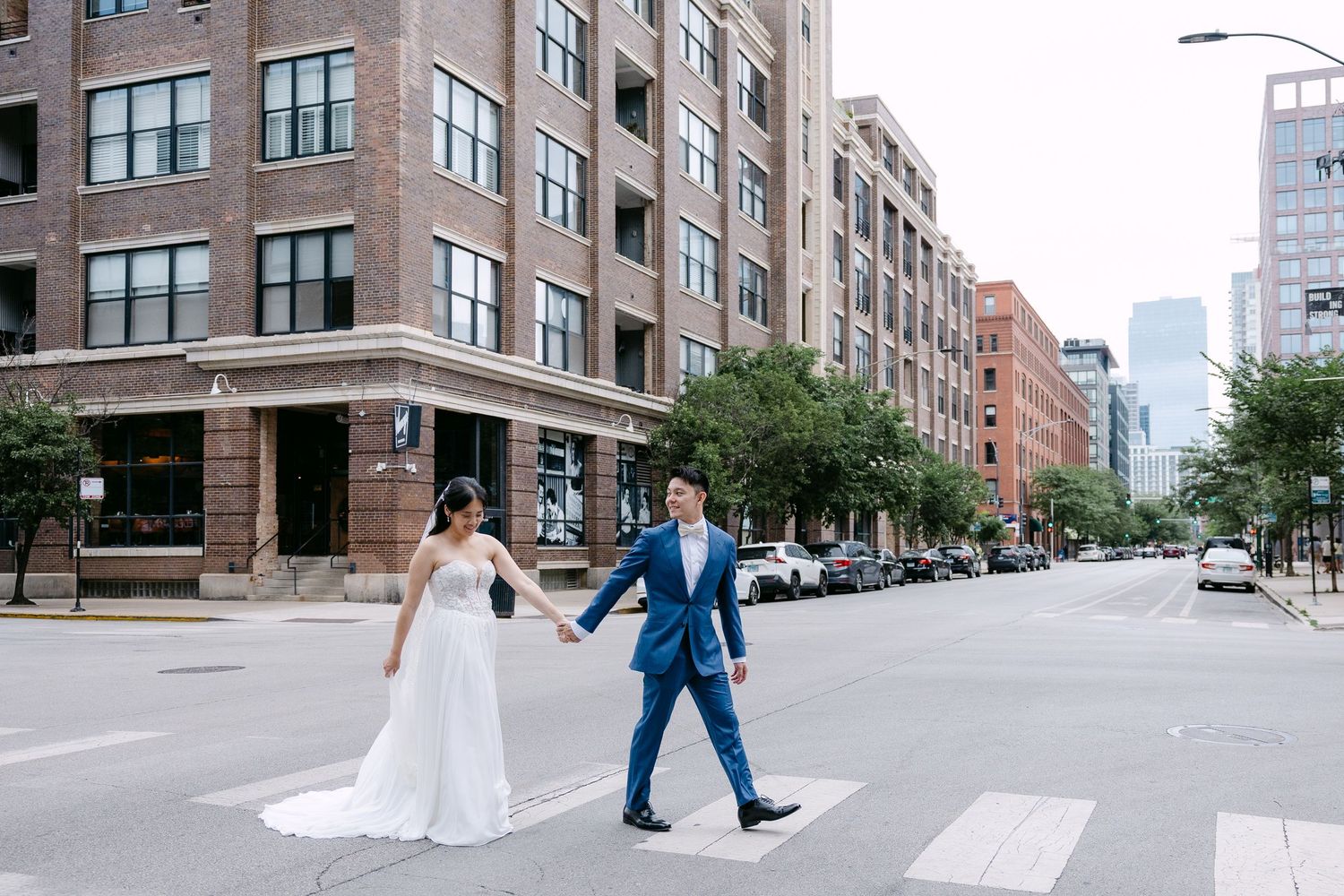 A couple in wedding attire crosses an urban street with brick apartment buildings and city skyline in the background.