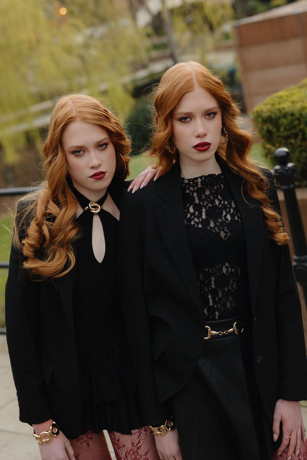 Two models in black lace dresses pose together against a garden backdrop with dramatic styling.