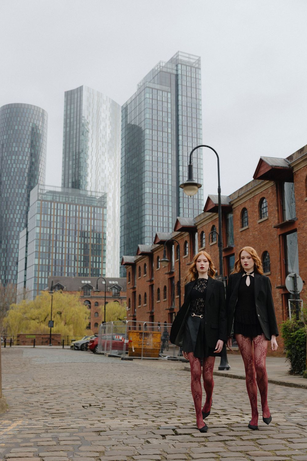 Two people walk along a cobblestone street with modern glass skyscrapers and historic brick buildings in the background.
