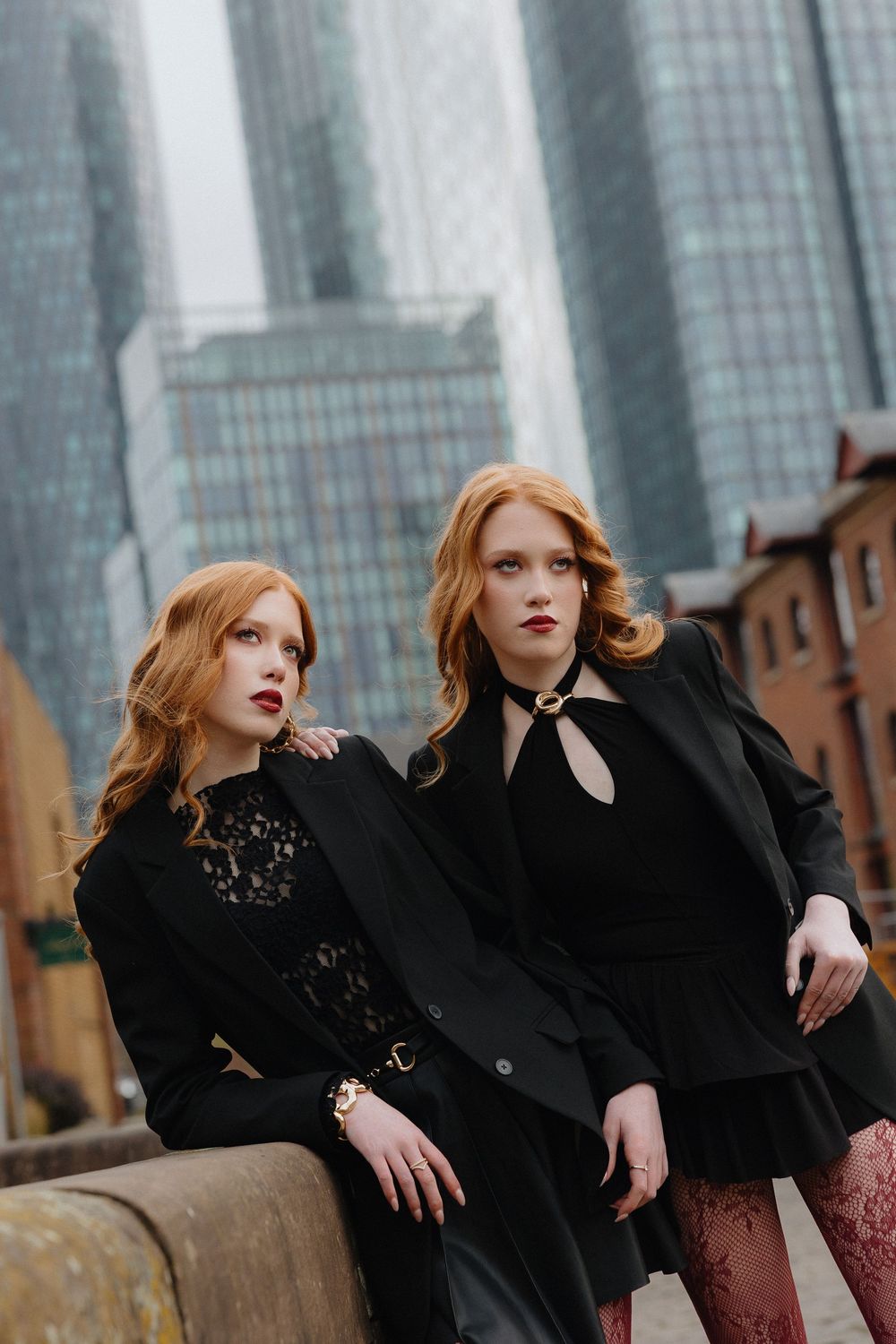 Two women in black gothic outfits pose dramatically against modern skyscrapers in an urban setting.