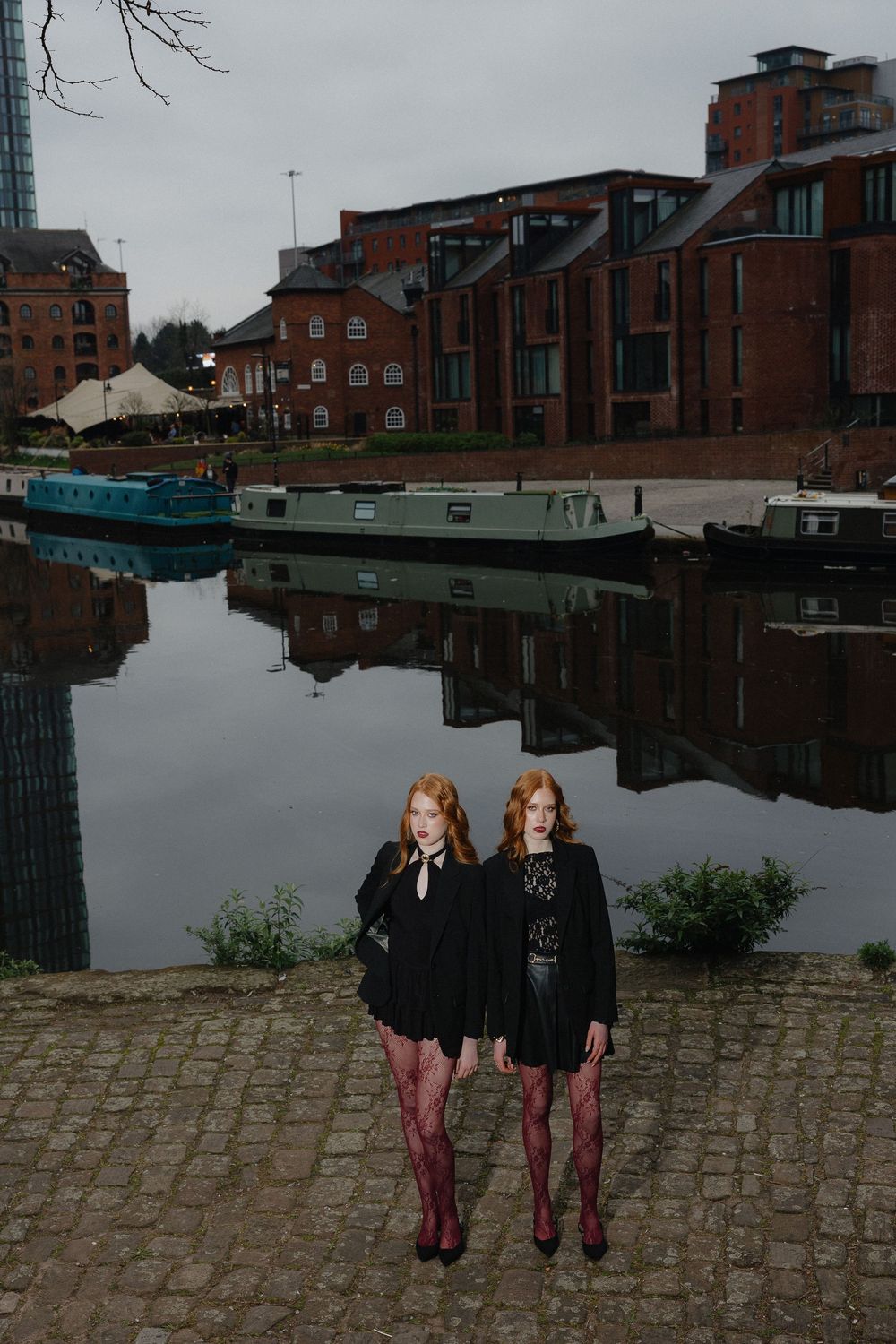 Two figures in black outfits with burgundy tights pose by a waterfront with brick buildings in the background.