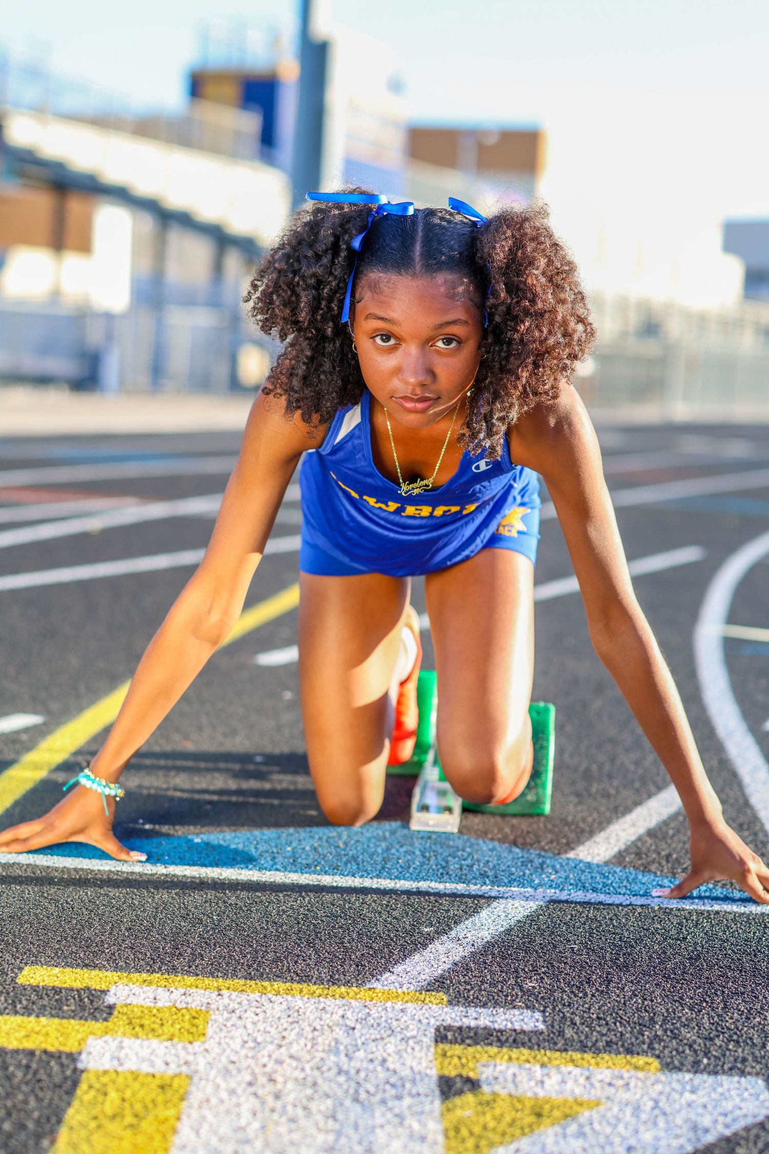 Track athlete in blue uniform crouches in starting position on running track with yellow and white lane markings.