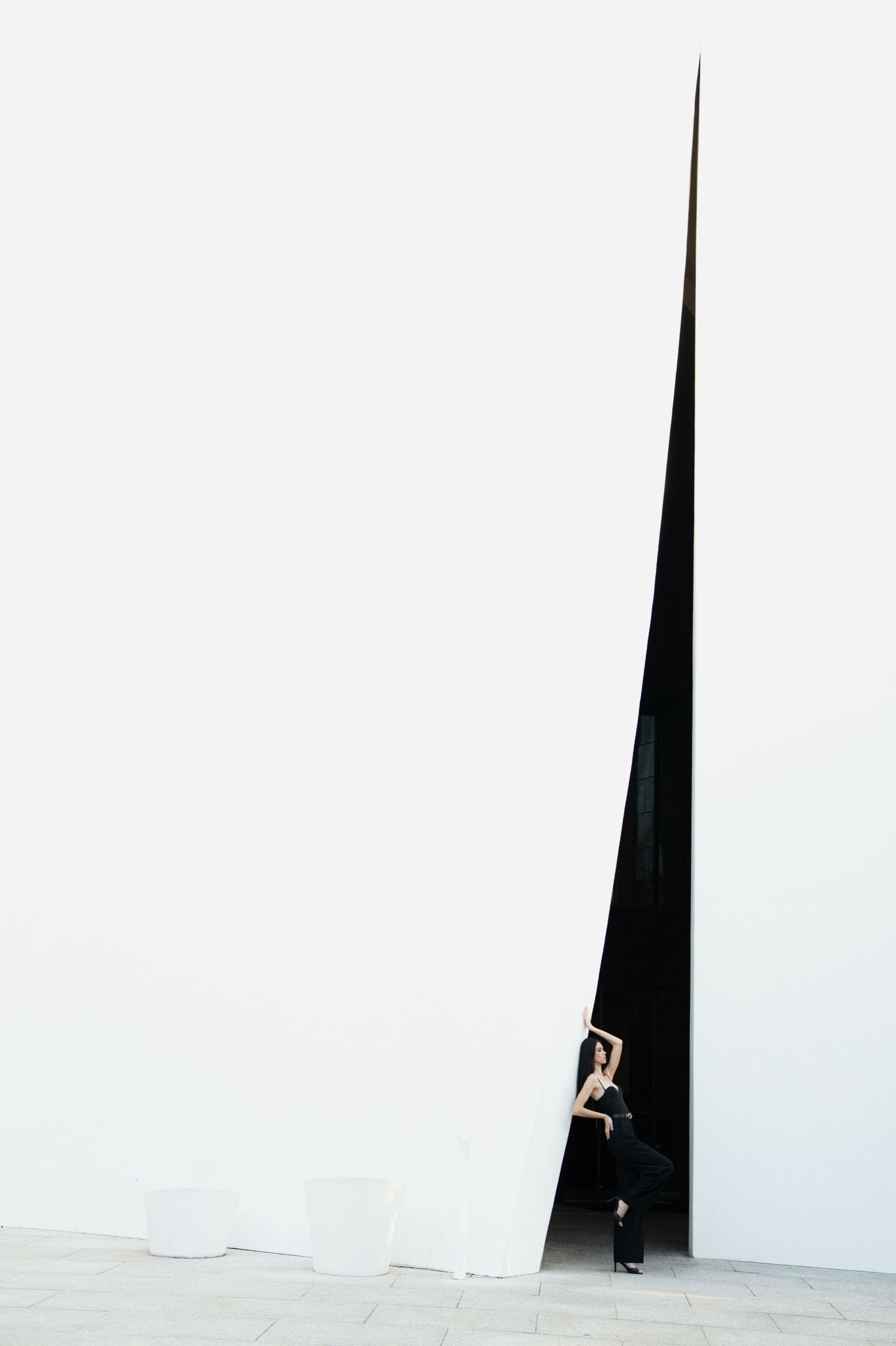 A person peeks through a dramatic triangular gap between stark white walls creating a minimalist architectural composition.