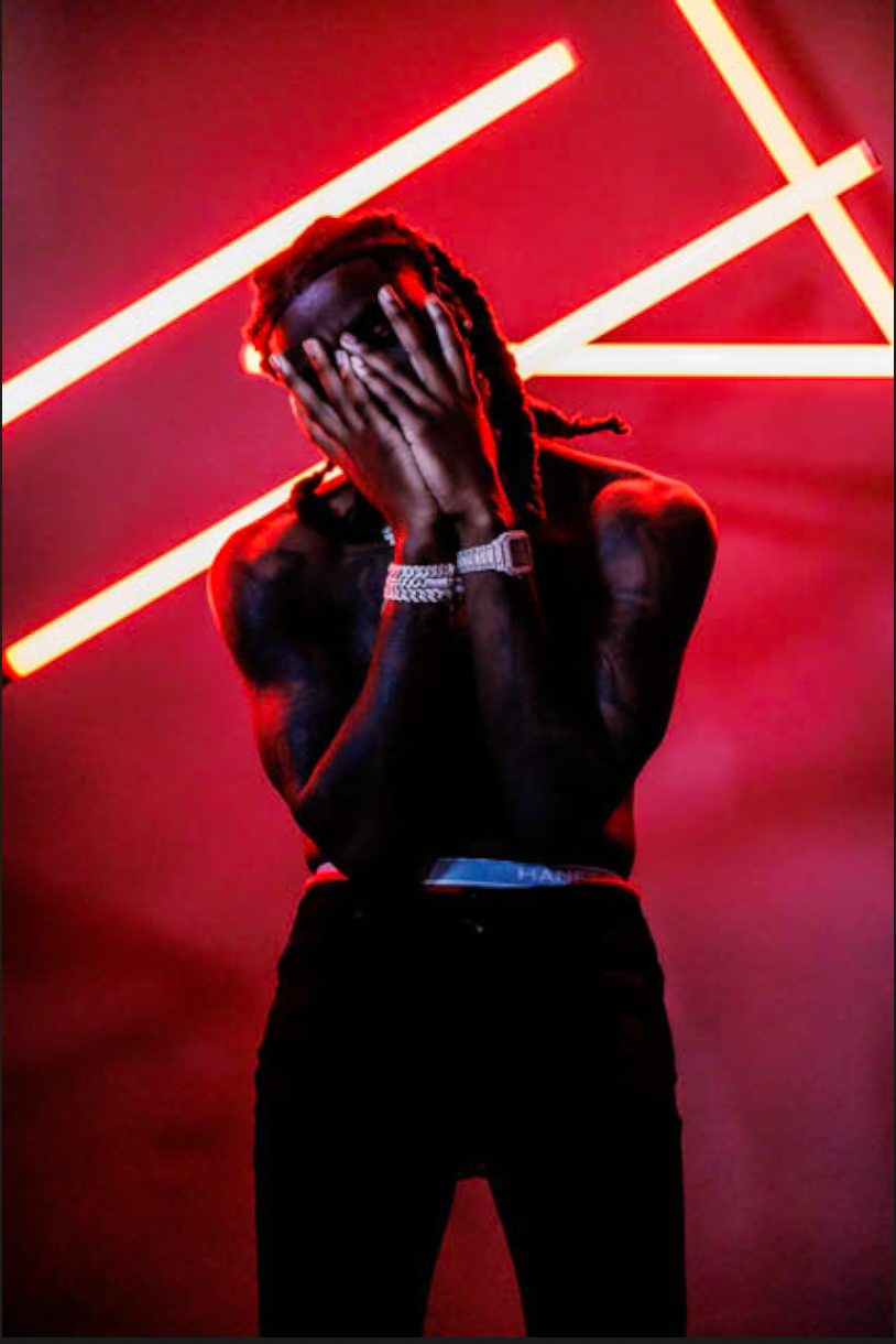 A shirtless Sleazy World Go covers his face with hands, wearing diamond bracelets, posed against red neon lights.