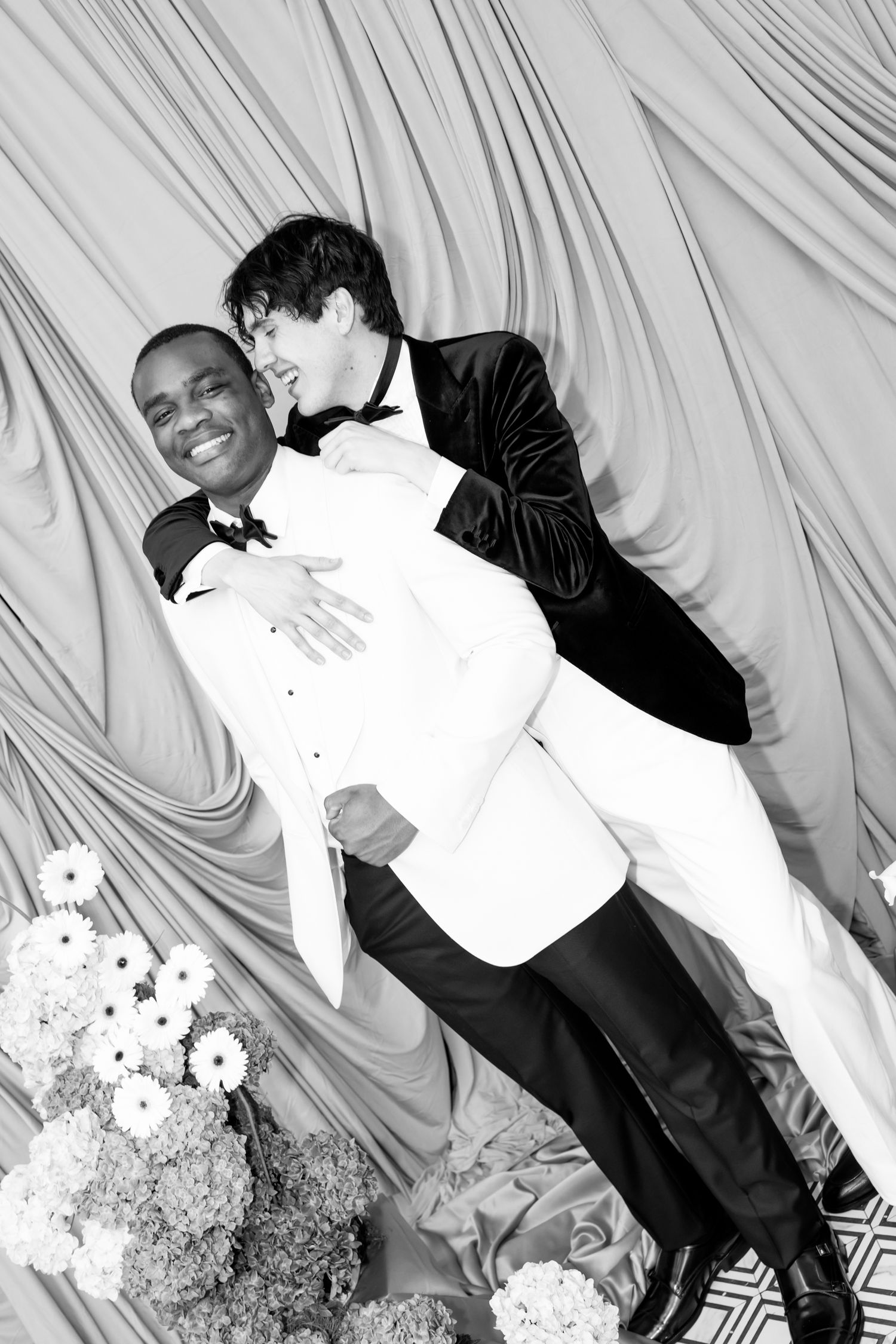 Two grooms in tuxedos embrace and laugh joyfully in front of elegant draped fabric at their wedding c at Bacchusmin Milwaukee