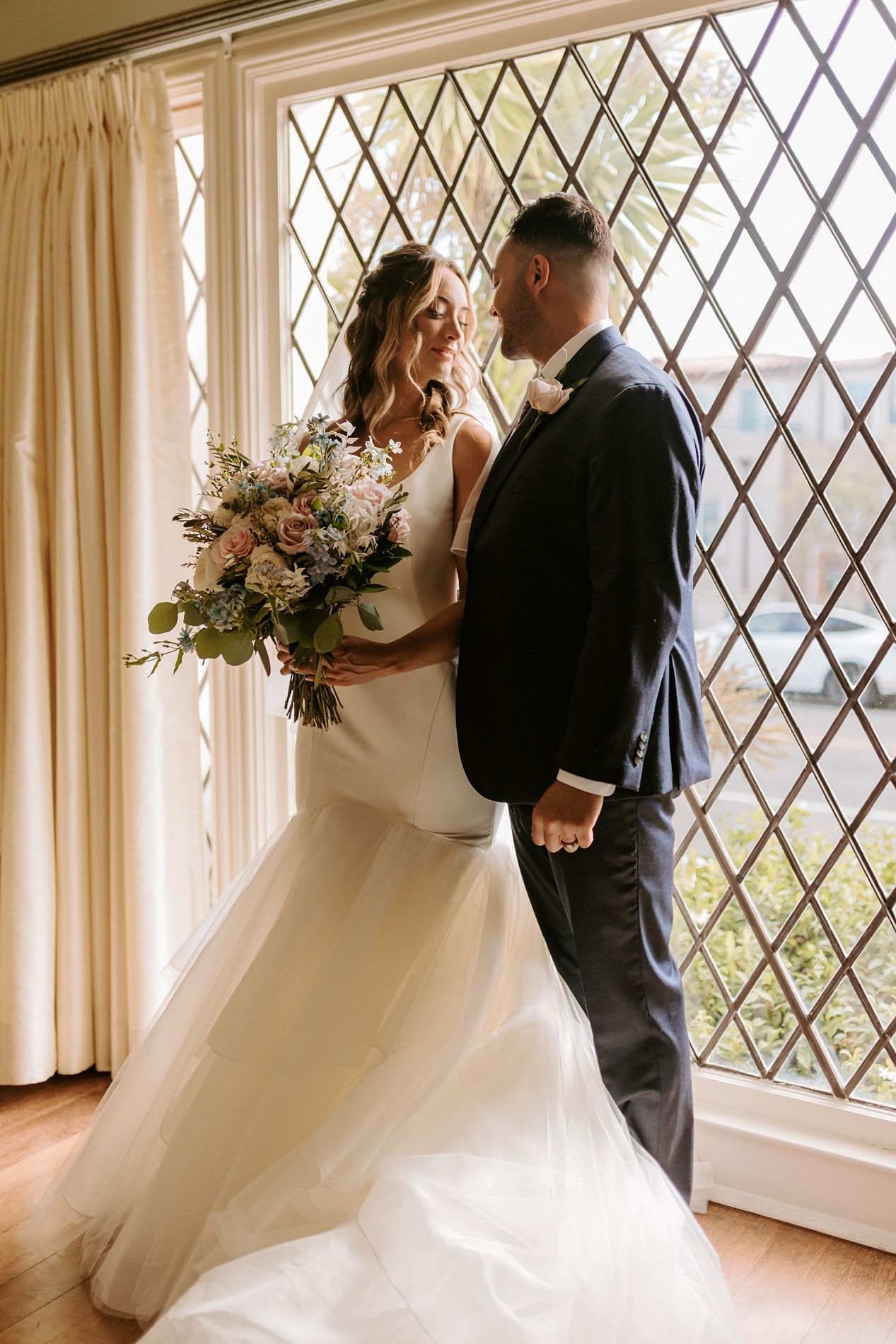 Bride and groom share a romantic moment by a leaded glass window, bride holding a lush floral bouquet in elegant wedding attire.
