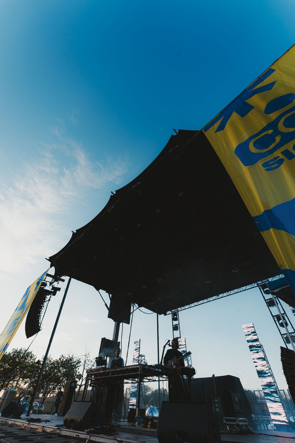 Silhouetted stage equipment and flags against a bright blue sky at an outdoor music venue.