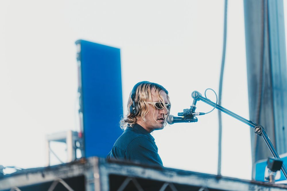 A musician performs behind a microphone on stage during an outdoor concert with a blue backdrop.