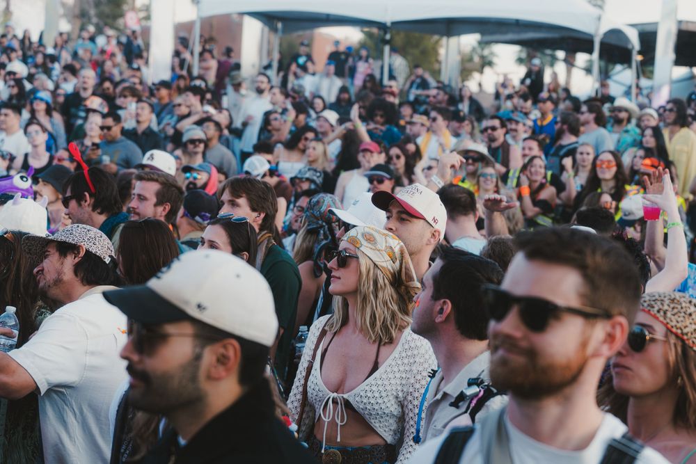 A large crowd of festival attendees wearing sunglasses and hats watches a performance during an outdoor event.