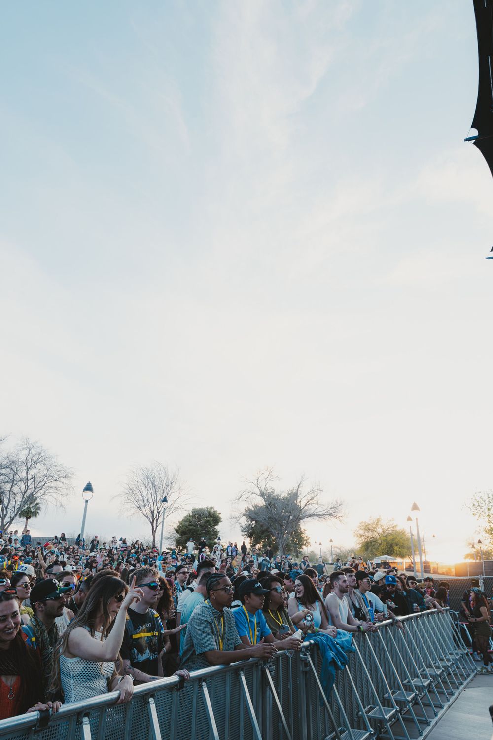 Large crowd of concert attendees stand behind metal barricades at an outdoor music festival during sunset.