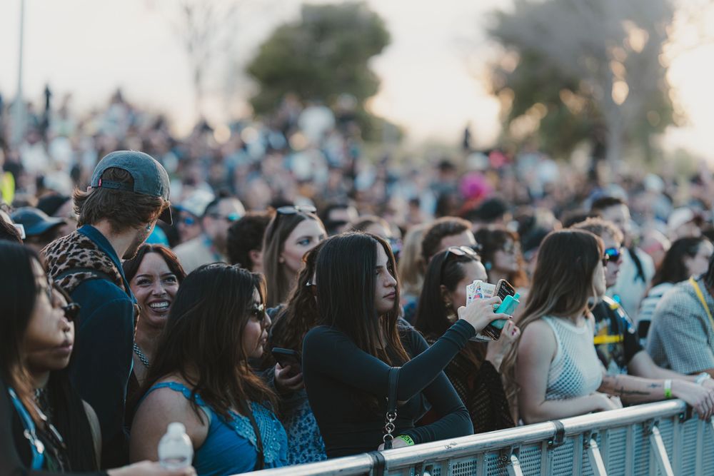 Large crowd of people enjoying an outdoor event with joyful expressions and raised hands in the sunlight.