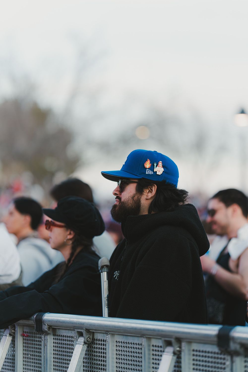 Baseball fans wearing dark jackets and blue caps watch from behind a railing at a stadium during a game.