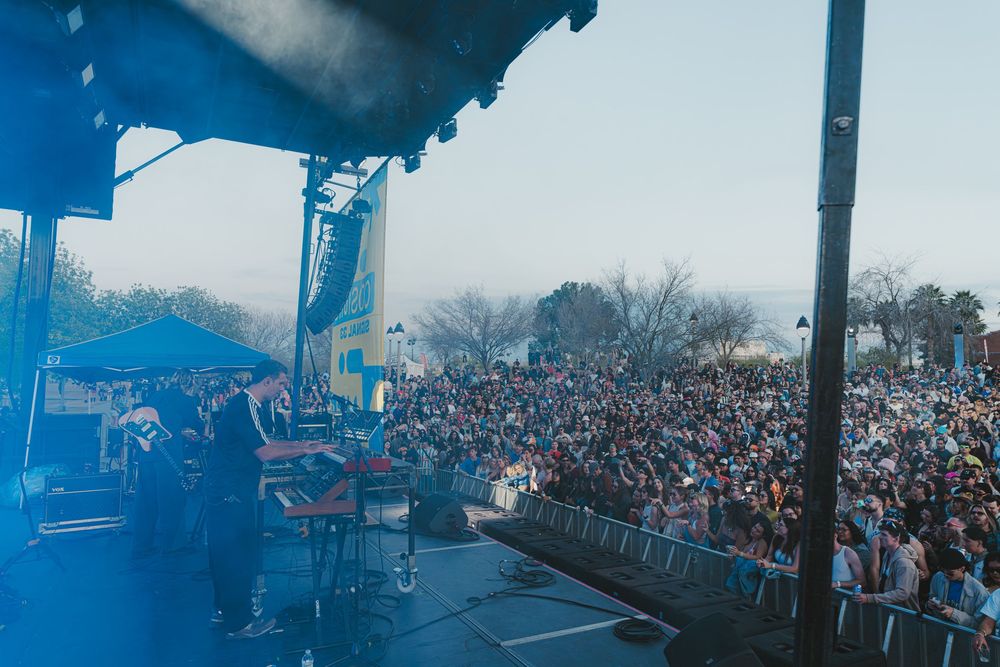 A large crowd of concert-goers fills an outdoor venue during a live music performance with blue lighting effects.