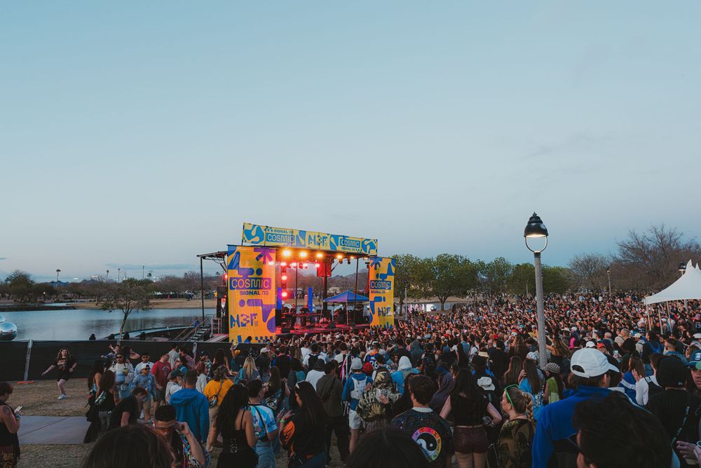 Large crowd gathered at an outdoor music festival with colorful illuminated stage during dusk.