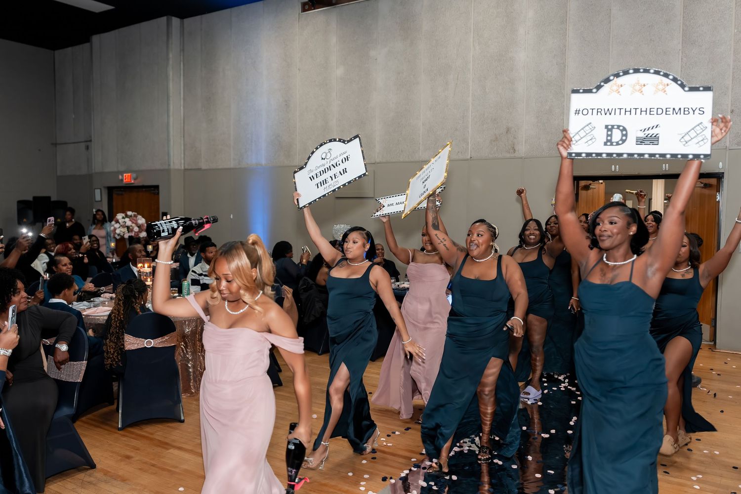 Wedding party in teal dresses dancing and holding signs during a celebration with confetti in the air.