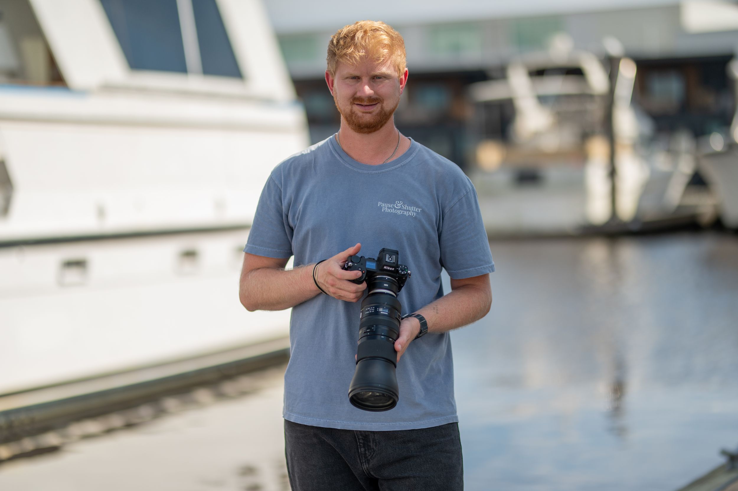 A photographer holding a camera stands near a marina with boats and buildings in the background.