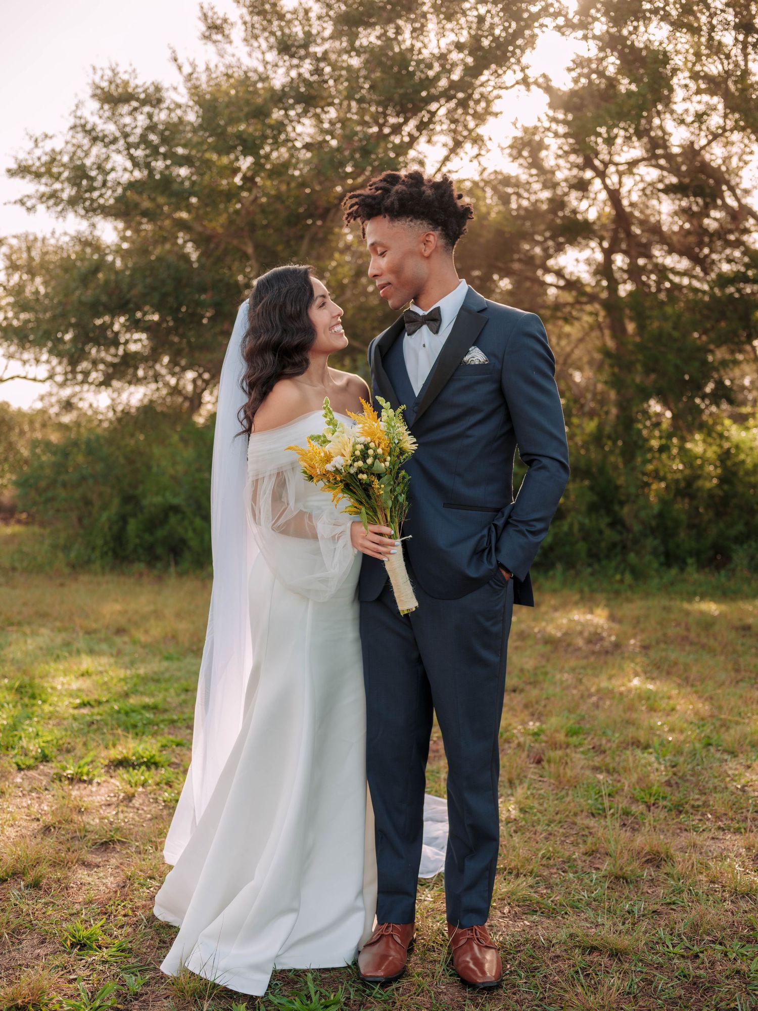 A wedding couple stands together outdoors at sunset, with the bride holding a yellow bouquet and groom wearing a navy suit.