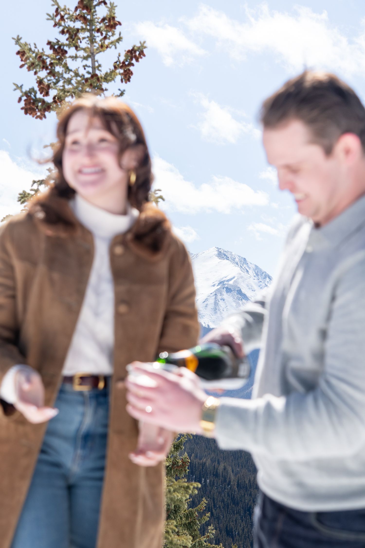 Two people enjoying drinks and laughter outdoors with snowy mountain peaks visible in the background for Aspen Colorado winter engagement.