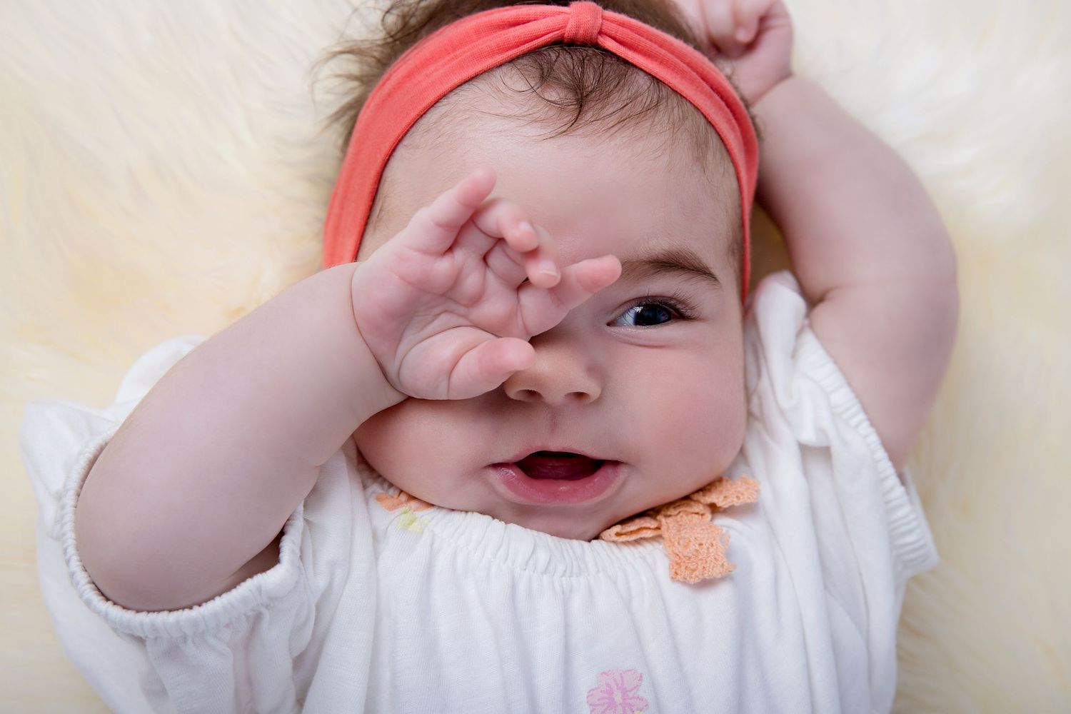 A smiling baby wearing a pink headband lies on a white blanket while reaching up with their hands.