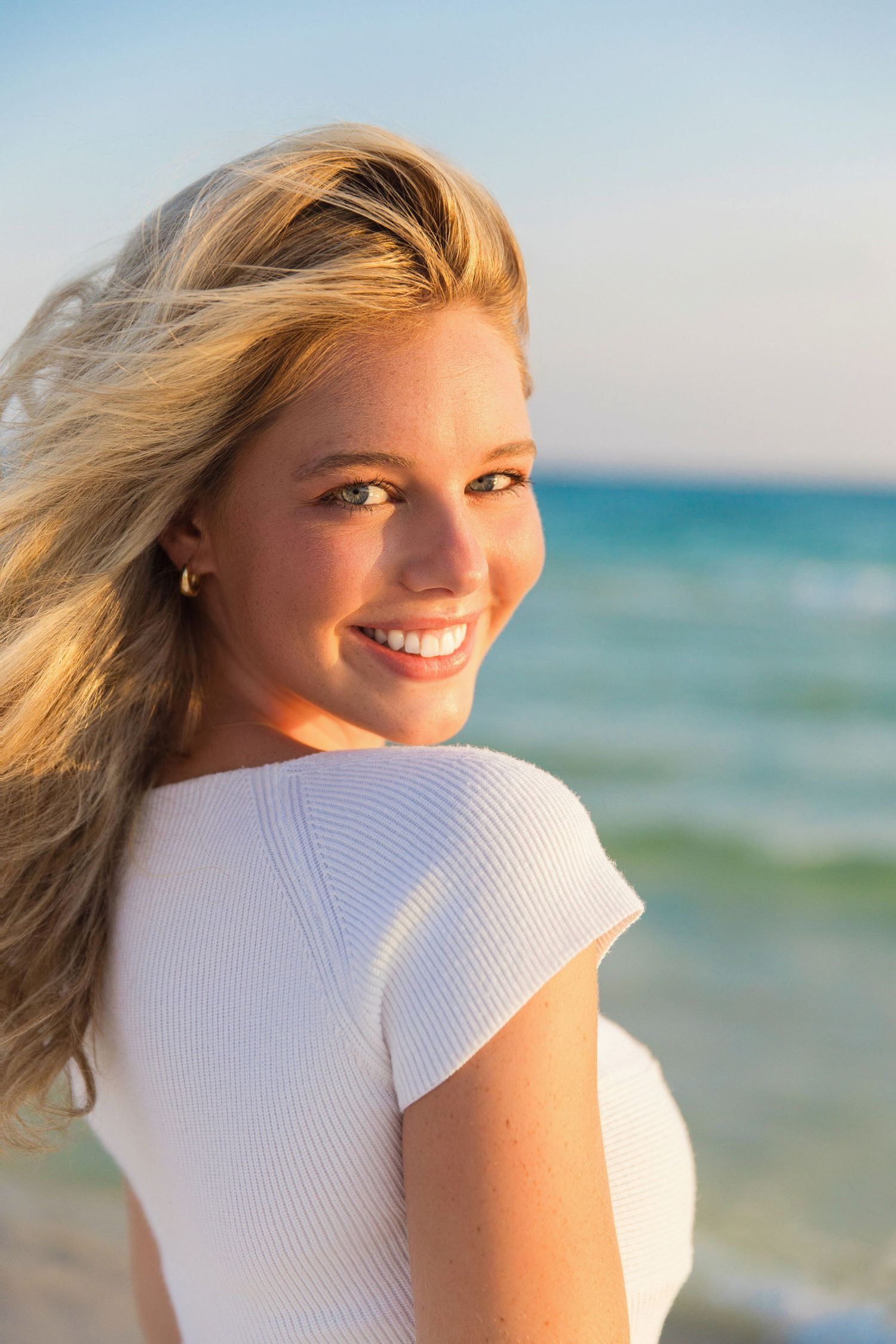 Woman in white top smiling on beach with ocean waves and blue sky in background.