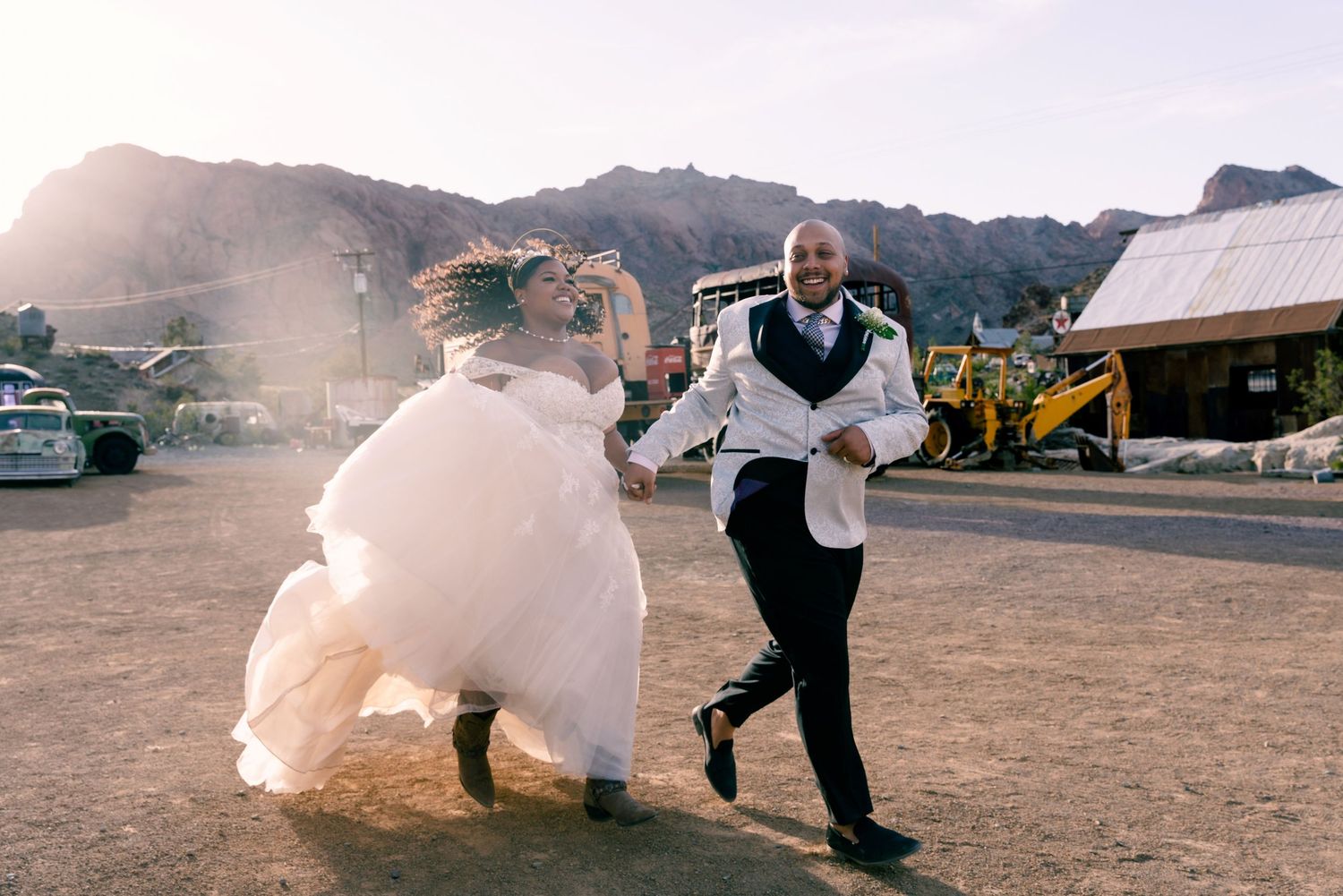 Two people run playfully across a dusty parking area with mountains in the background.
