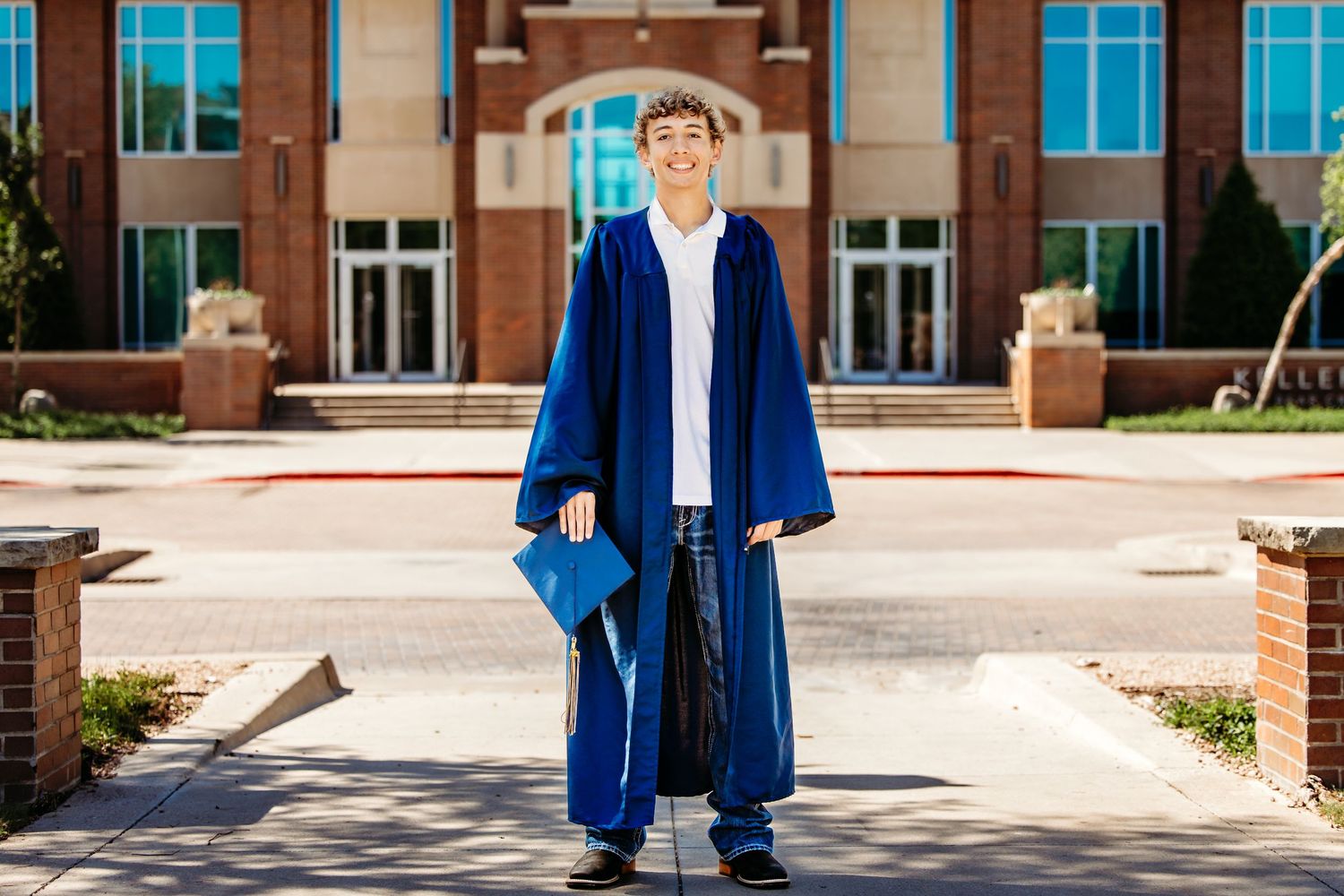 Young graduate in blue cap and gown standing proudly in front of Keller Town Hall, Keller, TX.