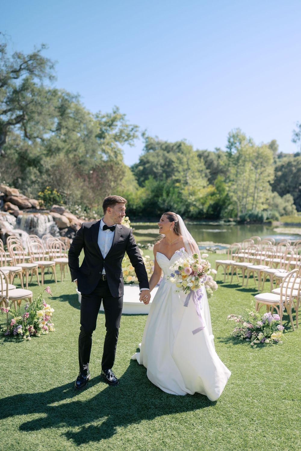 A couple walks hand in hand on their wedding day through an outdoor ceremony space with wooden chairs.