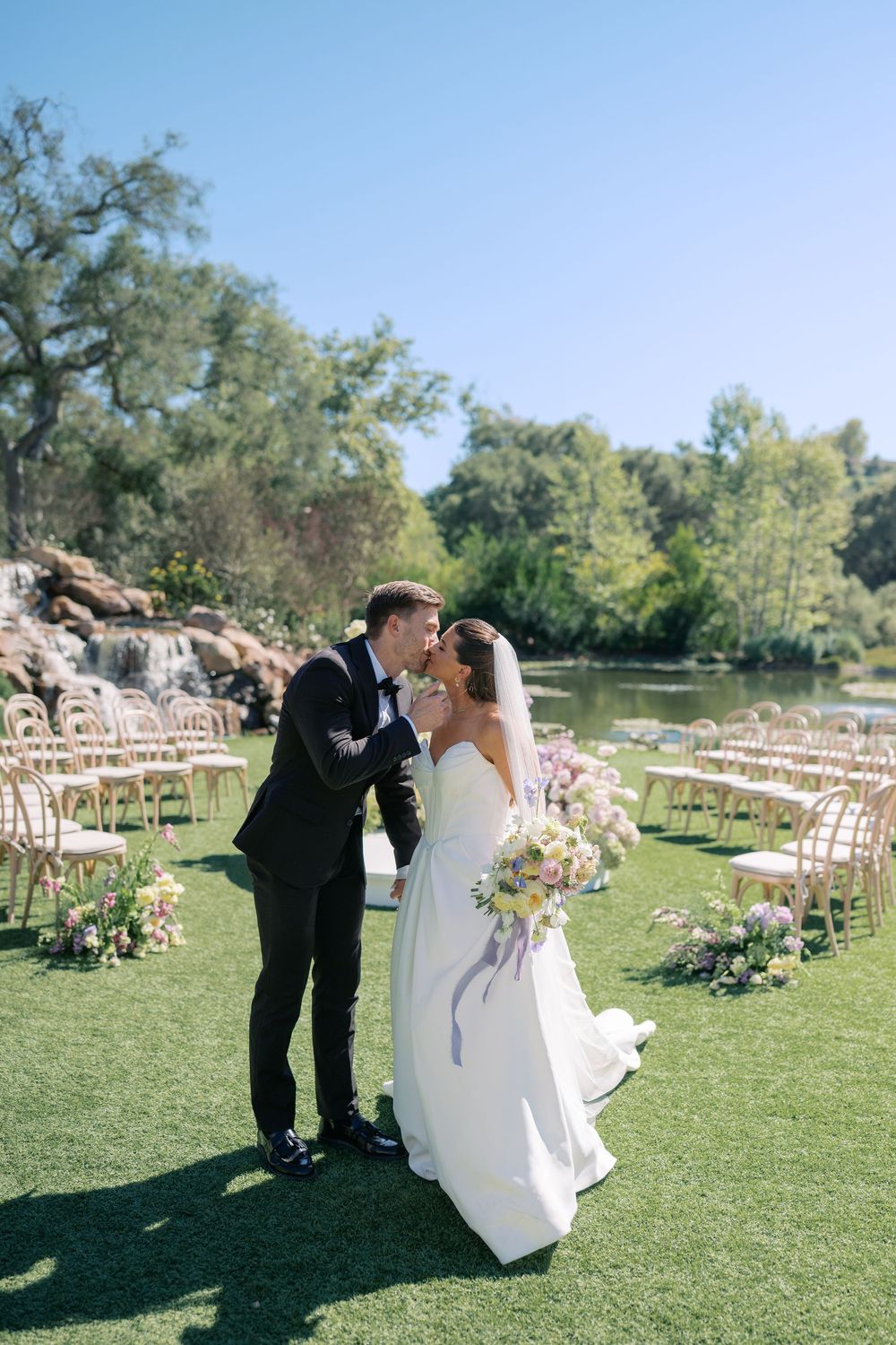 Couple in wedding attire share romantic moments on a sunny day at an outdoor ceremony with wooden chairs on green lawn.