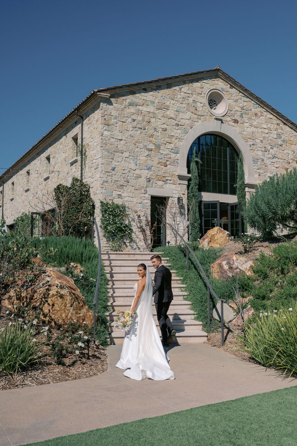 A couple stands outside a rustic stone building with arched windows and landscaped gardens on a sunny day.
