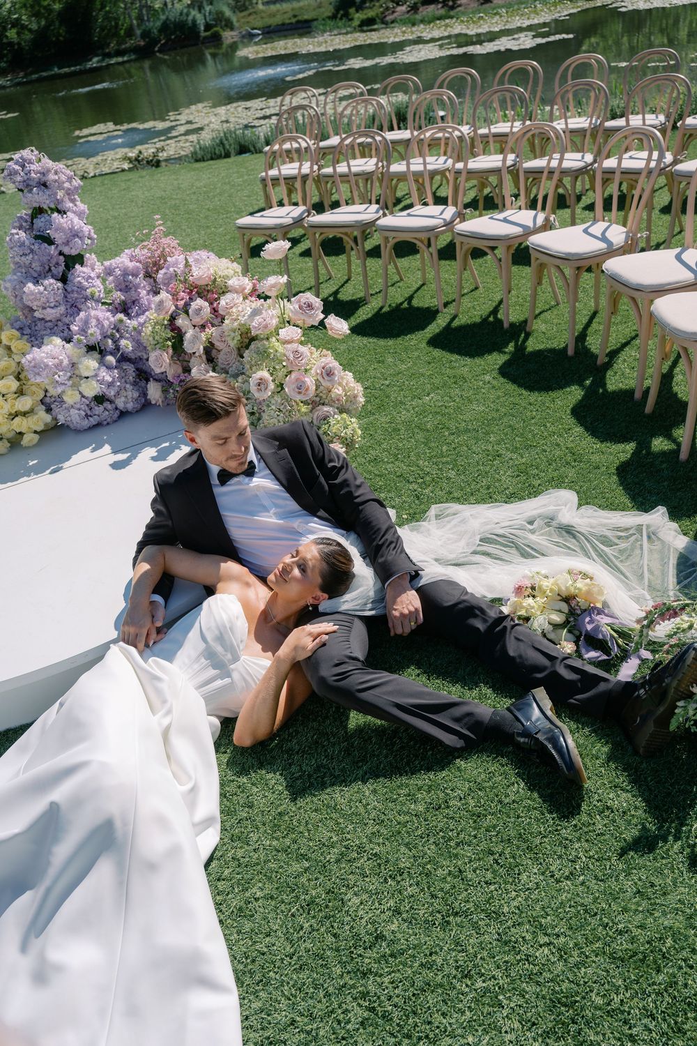 A romantic wedding couple poses playfully on manicured grass near elegant floral arrangements and ceremony seating.