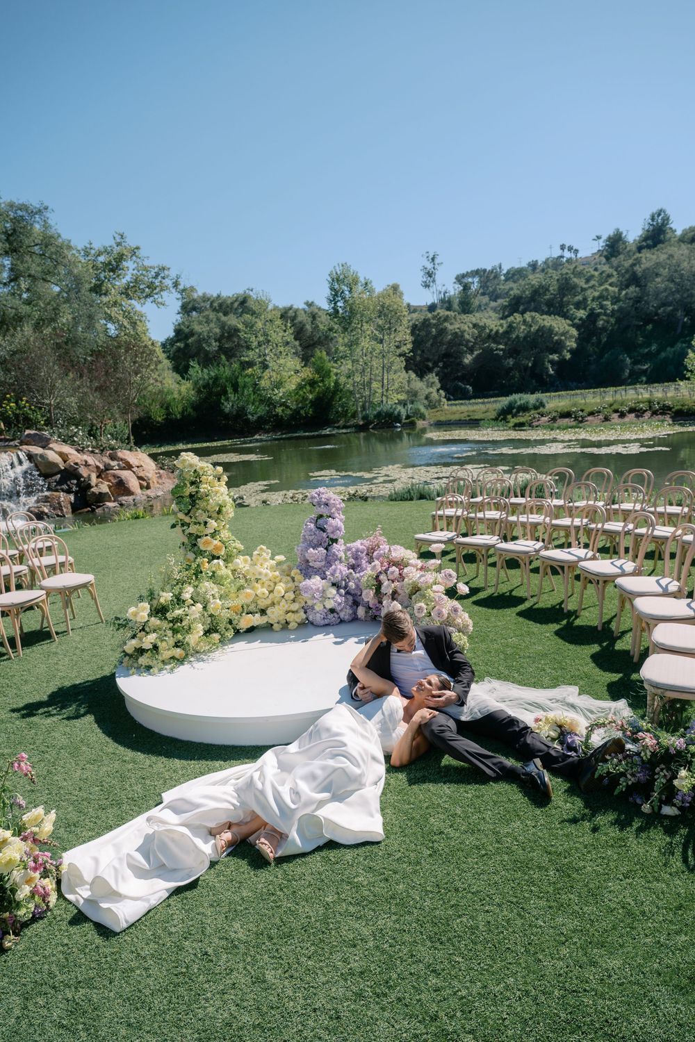 A wedding couple lies on a circular platform surrounded by floral arrangements and garden chairs.