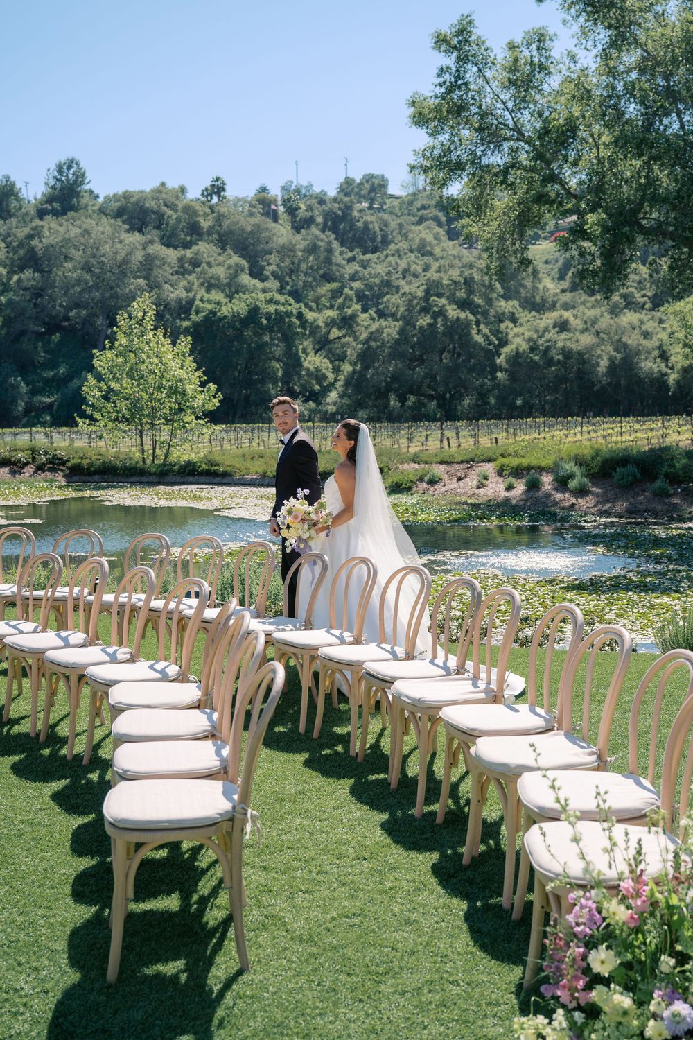 Rows of elegant wooden chairs with white cushions line a grassy outdoor wedding ceremony space beside a lake.
