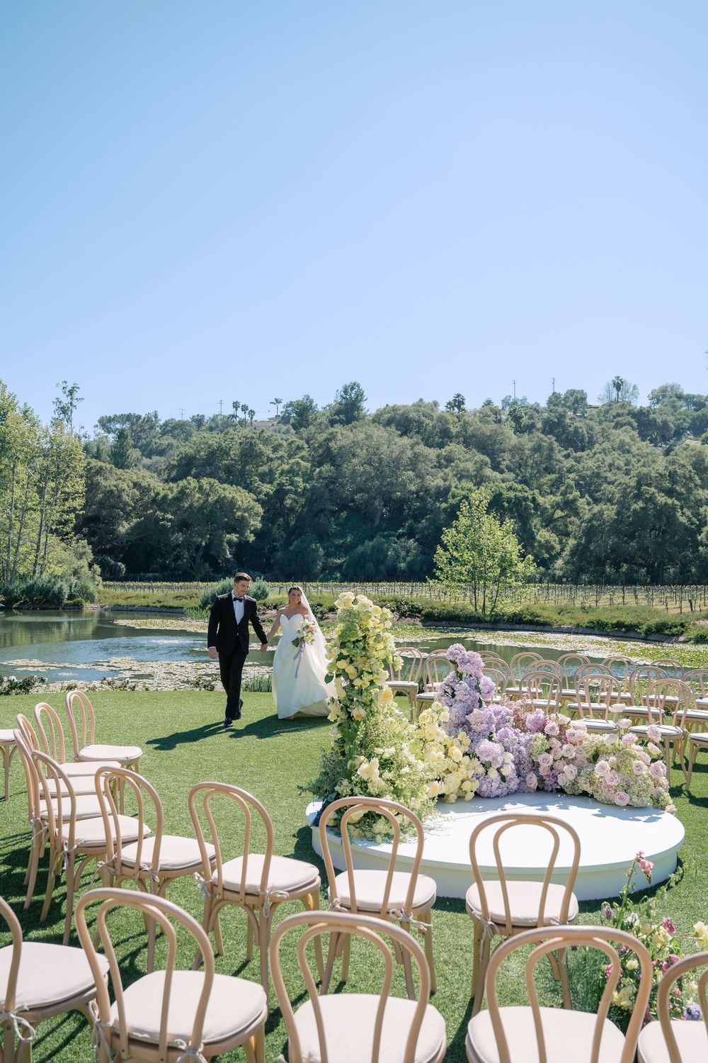 An outdoor wedding venue setup with wooden chairs and floral arrangements overlooking a scenic lake and wooded hillside.