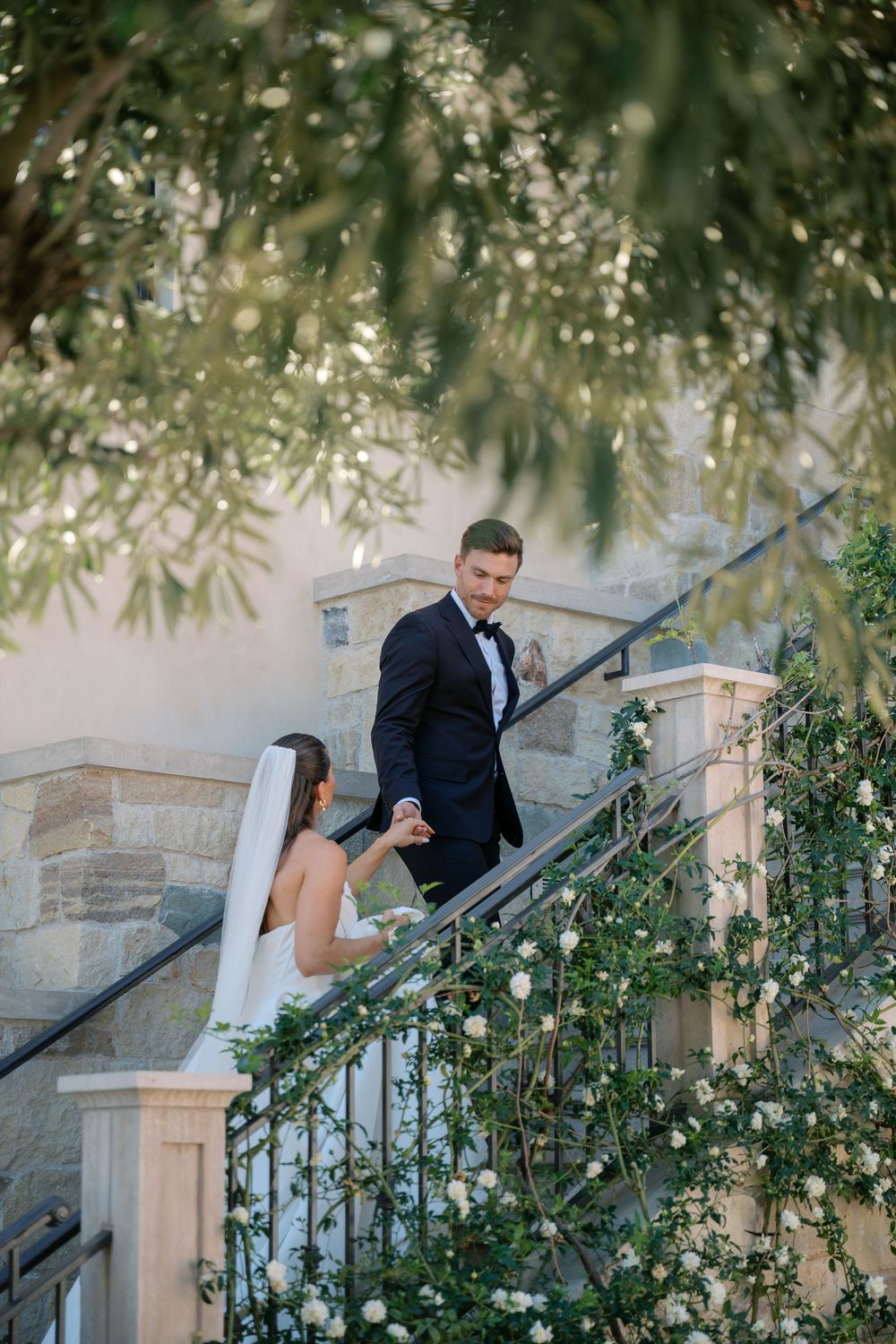 A groom in a black suit walks down stone steps surrounded by lush greenery and climbing vines.