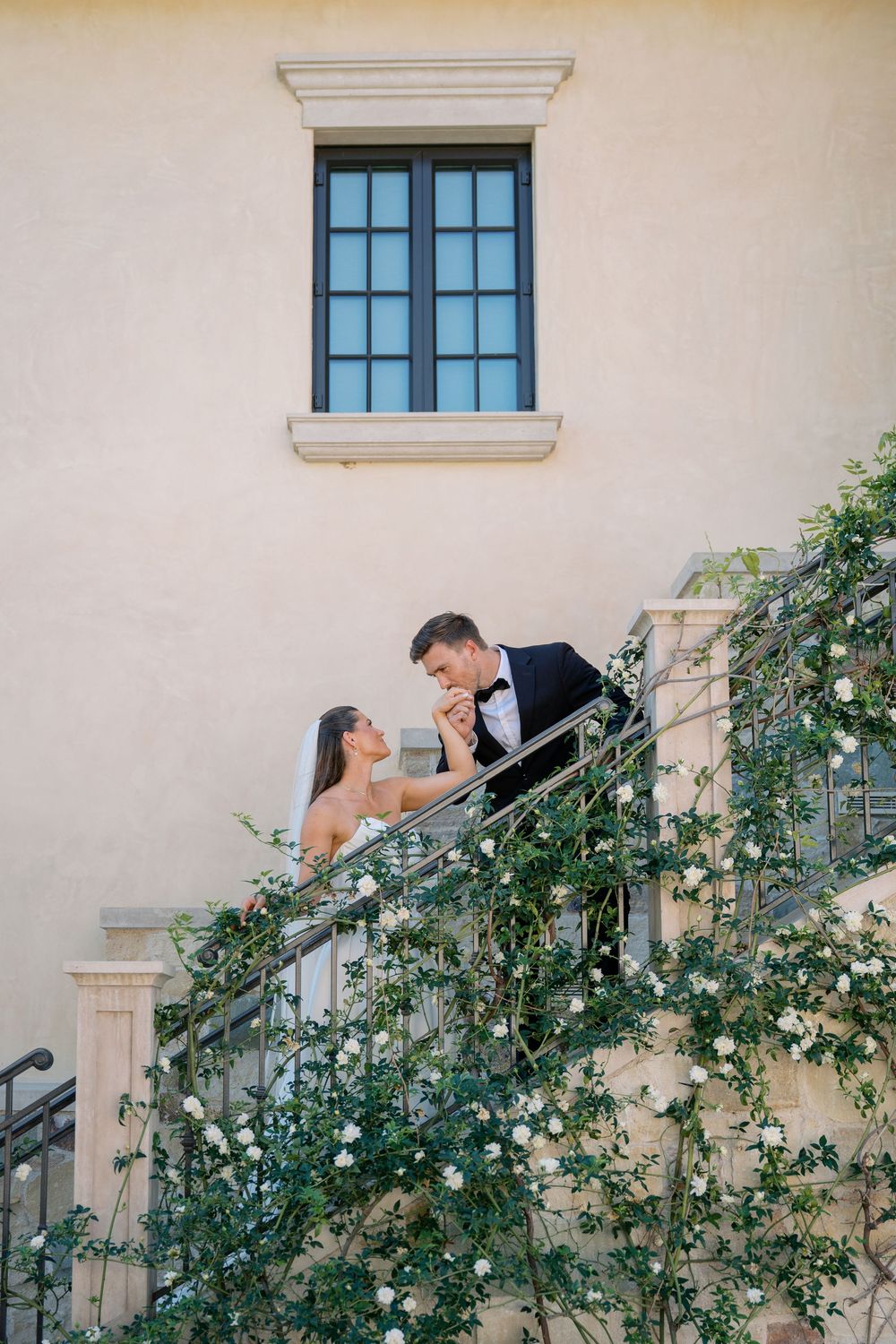 A romantic wedding moment captured against a cream colored wall with climbing roses and window details.