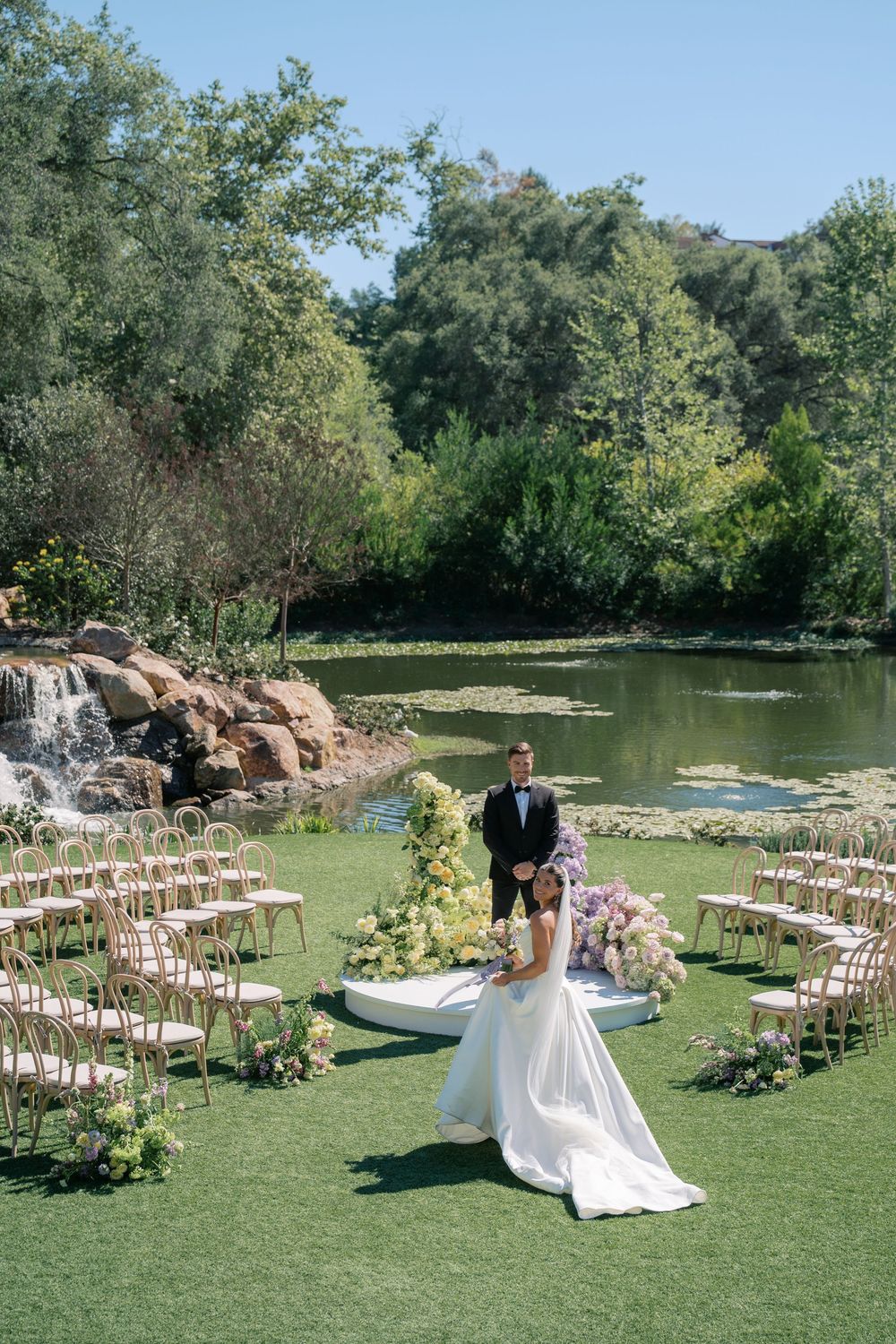 Outdoor wedding ceremony setup with white chairs arranged on green lawn beside a serene pond with waterfall feature.