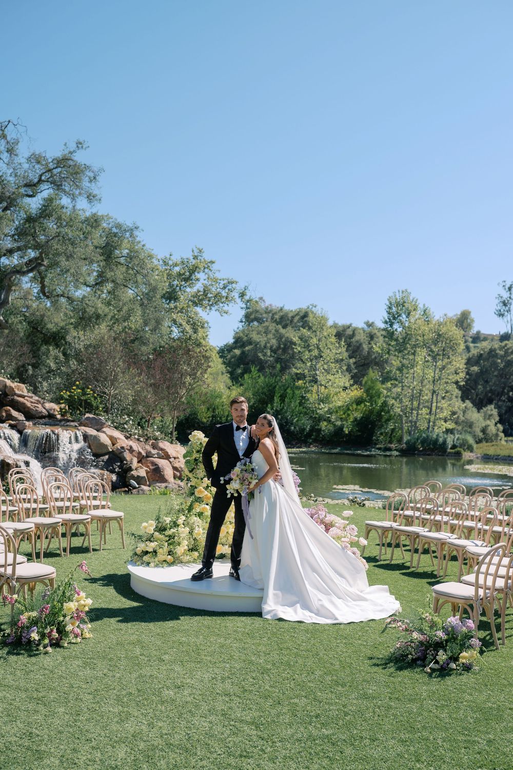 Outdoor wedding ceremony setup with wooden chairs arranged in rows on a lush green lawn beside a tranquil pond.