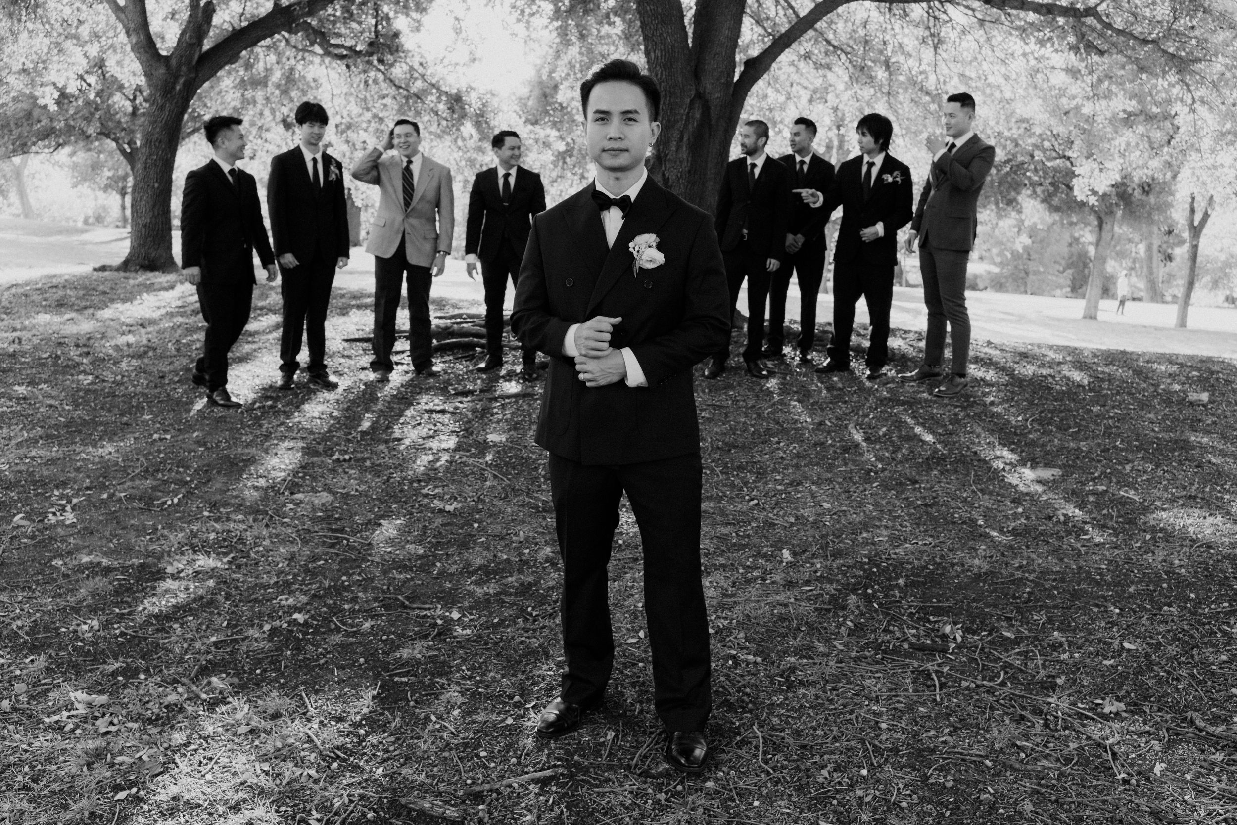 Black and white photo of groom in tuxedo standing before large wedding party group under shaded trees.