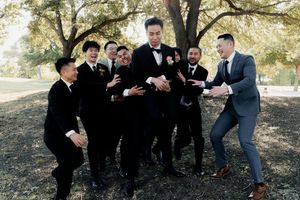 Groomsmen playfully surround groom in black tuxedo outdoors in a park setting with trees in background.