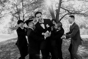 Black and white photo of laughing groom being playfully grabbed by groomsmen in sunlit outdoor setting.