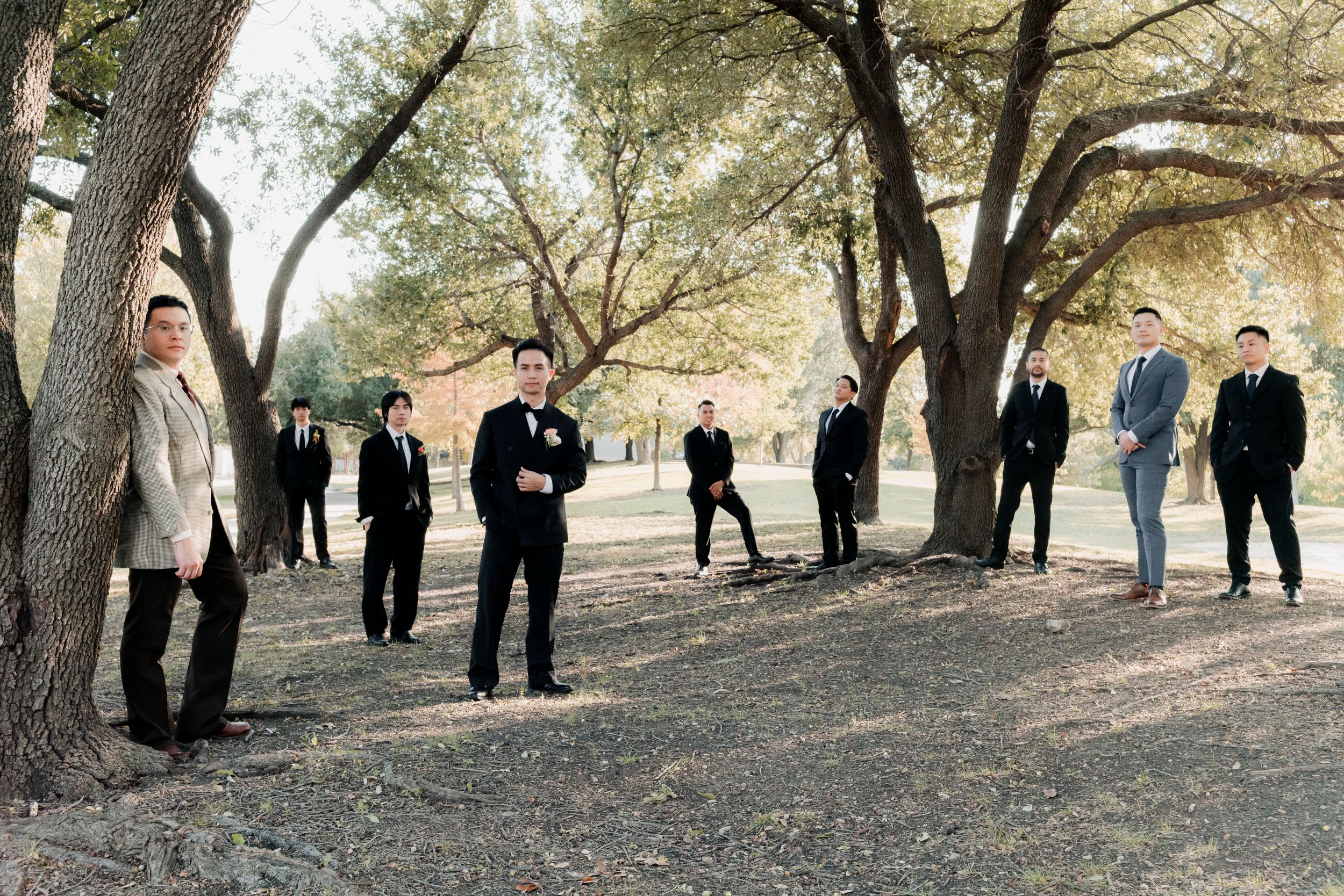 Groomsmen in suits posed individually among large trees in an outdoor park setting for wedding photos.