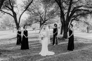 Black and white photo of bride in white gown with veil surrounded by bridesmaids in dark dresses outdoors.