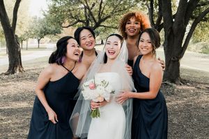 Bride in white strapless gown laughing joyfully with four bridesmaids in navy dresses outdoors among trees.