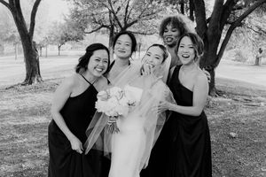 Bride with bridesmaids in black dresses posing joyfully outdoors in black and white wedding photo.