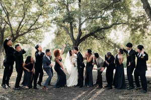 Wedding party celebrating under trees with bride and groom kissing surrounded by guests in black attire.