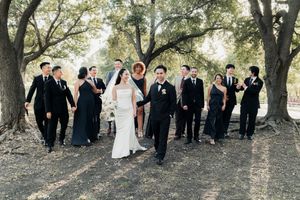 Bride and groom walk hand-in-hand with large wedding party through tree-lined outdoor venue.
