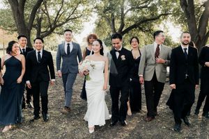 Bride and groom lead smiling wedding party walk through scenic outdoor venue with large trees.